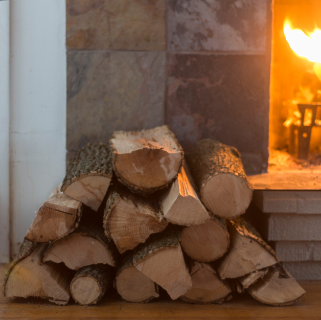 A stack of firewood sits next to a wood-burning fireplace, with flames burning brightly in the background.