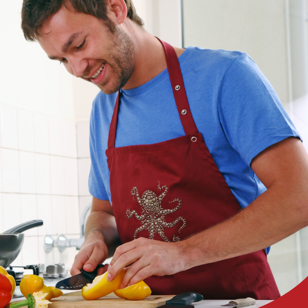 Man smiling while chopping yellow bell peppers in a kitchen with vegetables and a pan of food on the stove.
