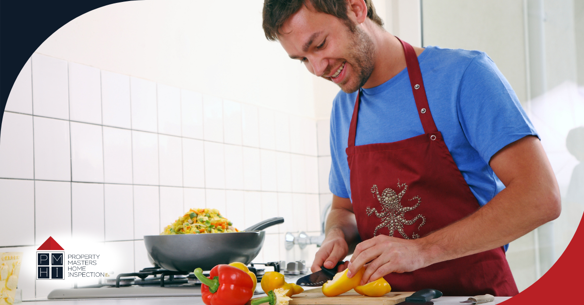 Man smiling while chopping yellow bell peppers in a kitchen with vegetables and a pan of food on the stove.